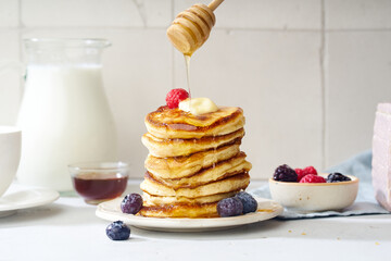 Pouring honey on a stack of fluffy breakfast pancakes with berries and butter