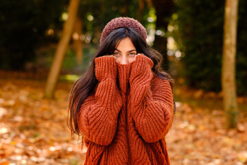 Young female in knitted hat covering face with warm sweater while standing in forest in cold autumn day