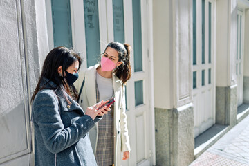Two women using their smartphones in the street with their face masks on.
