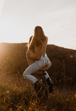 Unrecognizable shirtless boyfriend lifting topless girlfriend while standing in field at sunset and embracing gently