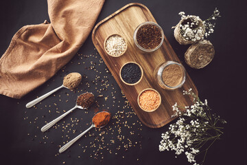Top view of assorted grains in jars and aromatic spices in spoons arranged on wooden tray on black table