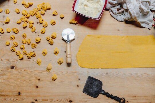 Top view of raw traditional tortellini scattered on wooden table in kitchen with wheel cutter and flatten dough