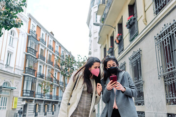 Two women using their smartphones in the street with their face masks on.
