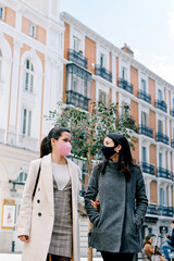 Two happy women talking and walking in the street with their face masks on.