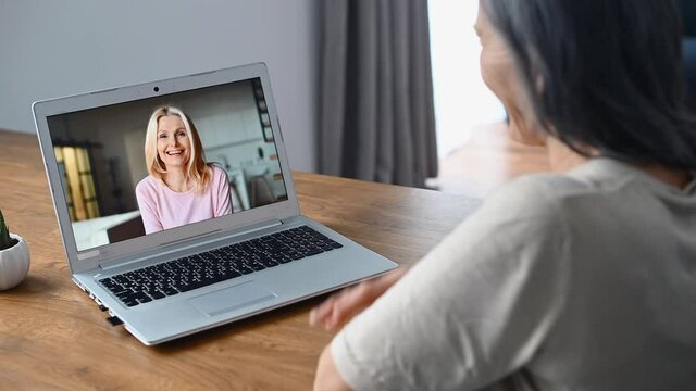 Two senior female friends has online meeting on the laptop, back view a senior woman talking via video call with her mate, employee, coworker, waving into webcam. Virtual conference concept