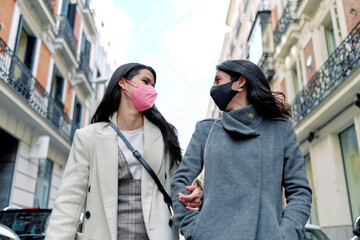 Two happy women talking and walking in the street with their masks on.