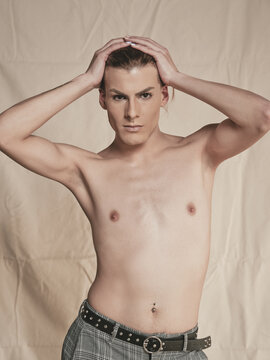 Shirtless androgynous guy looking at camera and touching head with manicured hand seductively against beige background