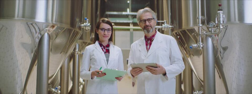 Colleagues in lab coat standing between rows of shiny stainless steel storage tanks. Looking at camera and smiling.