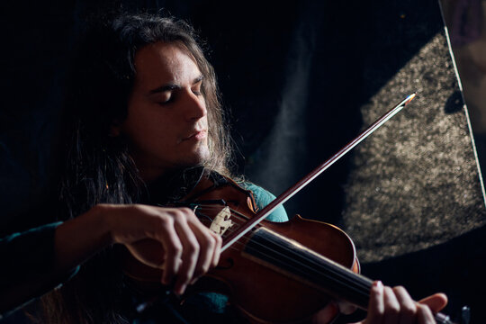 Talented male musician playing wooden violin with closed eyes while sitting in dark room