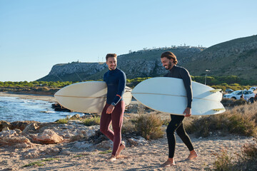 Positive male surfers in wetsuits and with surfboards walking along wet seashore after surfing at sunset