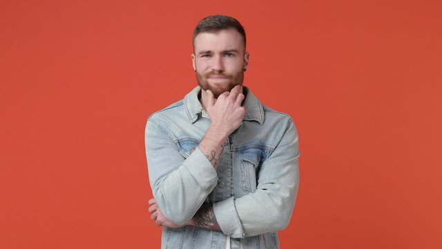 Confused puzzled bearded young man 20s years old in casual white t-shirt denim jacket isolated on orange color background in studio. People lifestyle concept. Put hand prop up on chin spreading hands