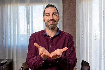 Young handsome bearded man wearing maroon shirt smiling with palms together receiving with palms together receiving or giving gesture