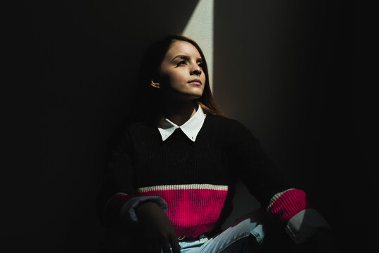 Serene female illuminated by bright sunbeam sitting in stylish clothes in dark room and looking up while showing concept of hope