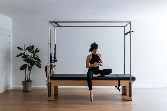 Fit female athlete sitting on pilates cadillac reformer and browsing cellphone while having break during training
