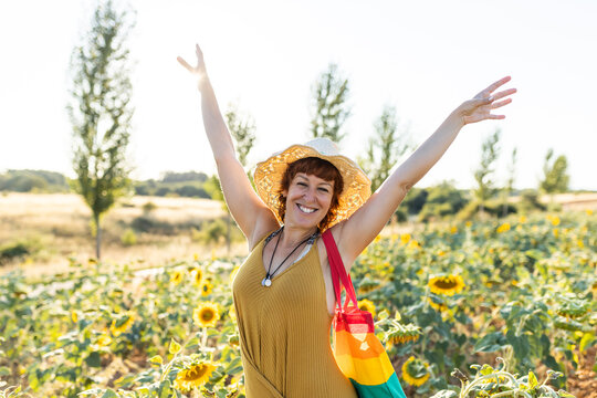Happy female with rainbow bag standing in booming sunflower field and enjoying weather and looking at camera