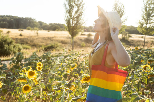 Side view of serene female with rainbow bag standing in booming sunflower field and enjoying weather