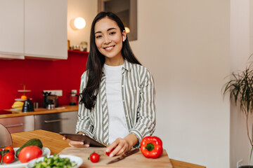 Lovely woman in striped shirt holding tomato and tablet. Smiling girl posing in kitchen