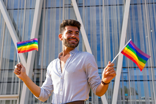 Low angle of delighted homosexual male standing on street with paper LGBT flags and looking away