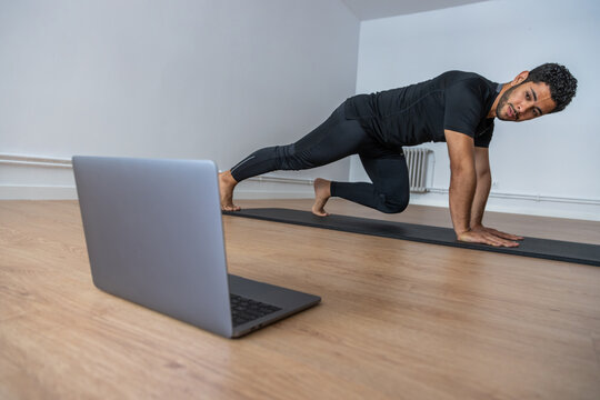Side view of concentrated ethnic male athlete practicing on mat during training while watching video tutorial on laptop