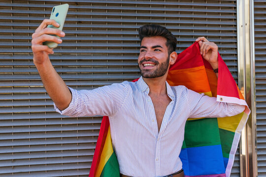 Content homosexual male taking self portrait with rainbow LGBT flag while using smartphone and standing near building in city