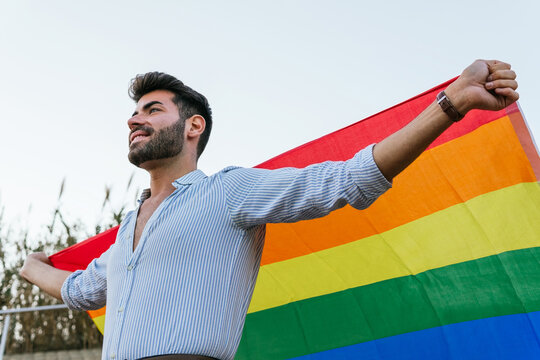 Low angle of expressive homosexual male standing on street with bright rainbow flag and cheerfully screaming