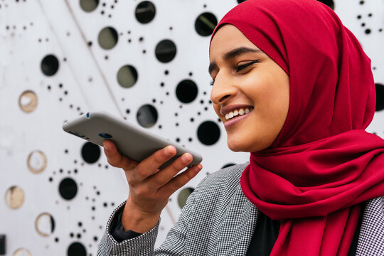 Side view of cheerful Arab female in hijab standing in street and recording voice message on mobile phone while smiling and communication on social media