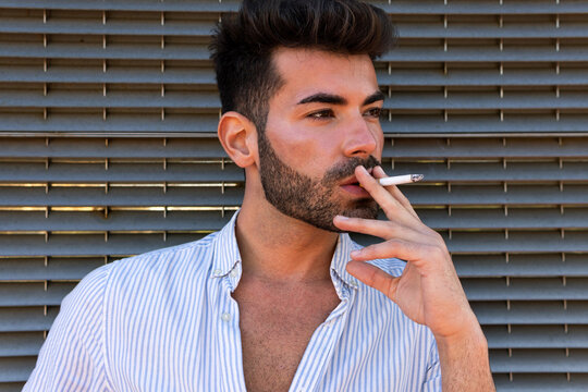 Thoughtful handsome male leaning on building and smoking cigarette in city while looking away in contemplation