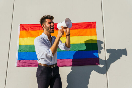 Side view of excited homosexual male standing near building with LGBT rainbow flag and shouting in loudspeaker
