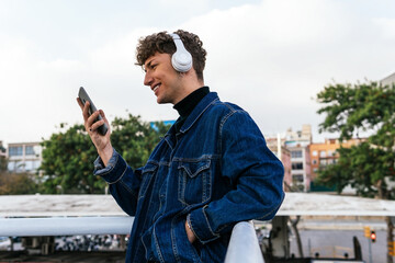 Side view of positive young male listening to music in wireless headphones browsing on smartphone while enjoying weekend in street