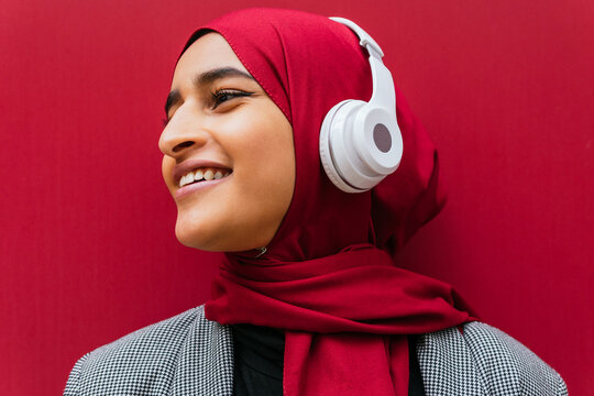 Side view of dreamy Arab female in wireless headphones and traditional hijab standing near urban red building and enjoying music looking away