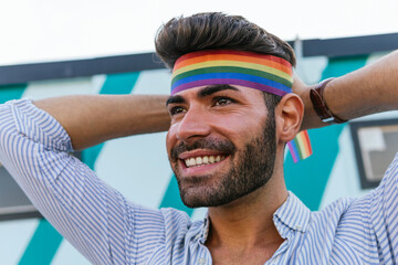 Low angle of homosexual male putting on bright headband in colors of rainbow LGBT flag while standing in city and looking away with smile