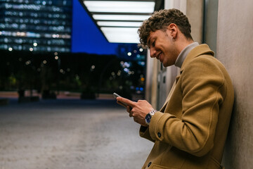 Side view of smiling handsome male leaning on building and messaging on social media via smartphone in evening in city
