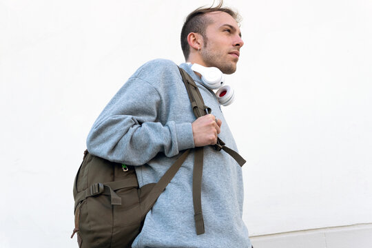 From below side view of modern young hipster male in casual outfit with backpack walking near light stone wall on urban street