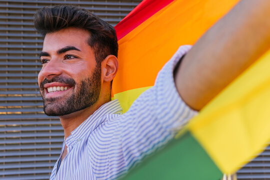 Positive gay male standing with rainbow LGBT flag in city and looking away