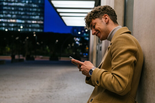 Side view of smiling handsome male leaning on building and messaging on social media via smartphone in evening in city