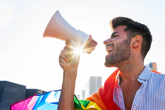 Side view of eccentric gay male with rainbow LGBT flag standing in city and yelling in loudspeaker with closed eyes