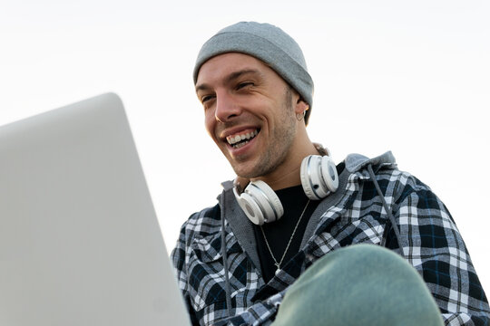 Low angle of happy young hipster male blogger in casual outfit with headphones on neck sitting on stone border and browsing laptop at sunset time