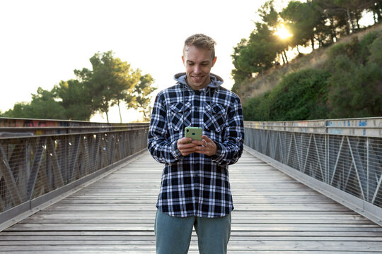 Positive young male traveler in checkered shirt checking data on smartphone while standing on wooden footbridge during summer journey in countryside