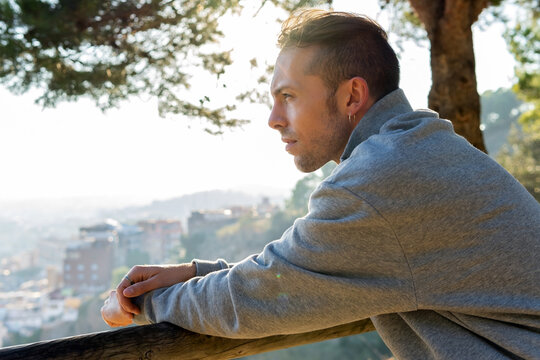 Side view of pensive dreaming young hipster male in casual outfit leaning on wooden railing and looking away while standing on green hill against blurred city