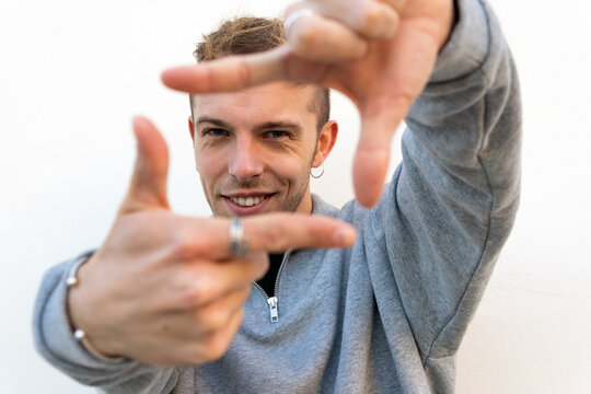 Smiling young hipster guy in casual outfit making photo frame with fingers and looking at camera against white background