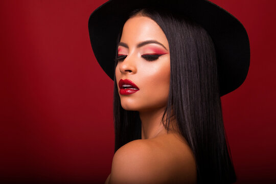 Magnificent female with red lips and in black hat standing with opened mouth and closed eyes on red background in studio