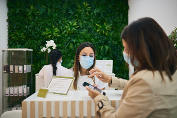 Female receptionist in mask standing at counter in modern beauty salon and waiting for payment from woman for skin care procedure