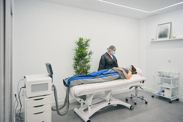 Woman in costume for pressure therapy lying on medical table and talking to beautician in mask in contemporary beauty clinic