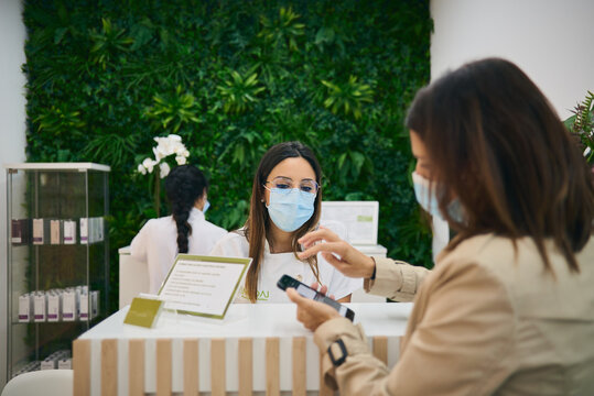 Female receptionist in mask standing at counter in modern beauty salon and waiting for payment from woman for skin care procedure