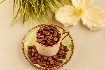 Roasted brown coffee beans poured into beautiful ceramic coffee cup, white flower and green grass on light background. Coffee still life. Flat lay, top view.