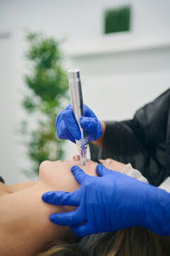 Cropped unrecognizable cosmetologist in uniform using derma pen with micro needles and cleaning face of client in beauty clinic