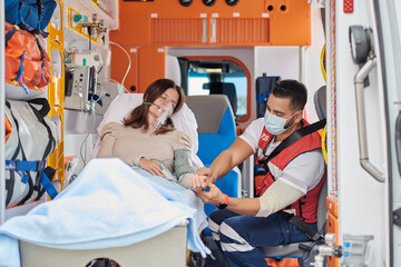 Paramedic putting finger pulse oximeter on patient in ventilator mask lying on medical stretcher in ambulance car