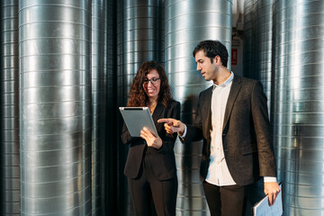 Busy male and female colleagues in formal clothes using tablet together while working in storehouse with metal barrels
