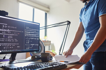 Crop unrecognizable male physics student standing at table with documents and computer displaying various formulas