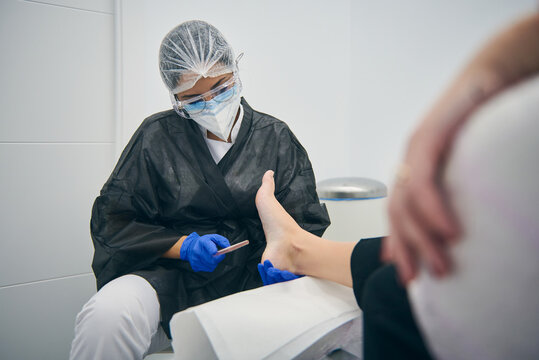 Female podologist in uniform doing pedicure for woman sitting in medical chair in beauty clinic